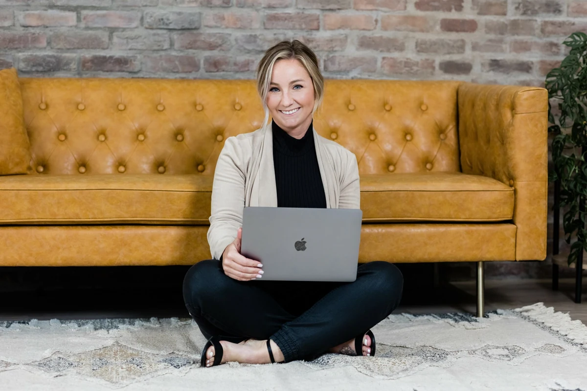 Randi Van Horn sits cross-legged on a rug in front of a tan leather sofa, smiling, with a laptop on her knees.