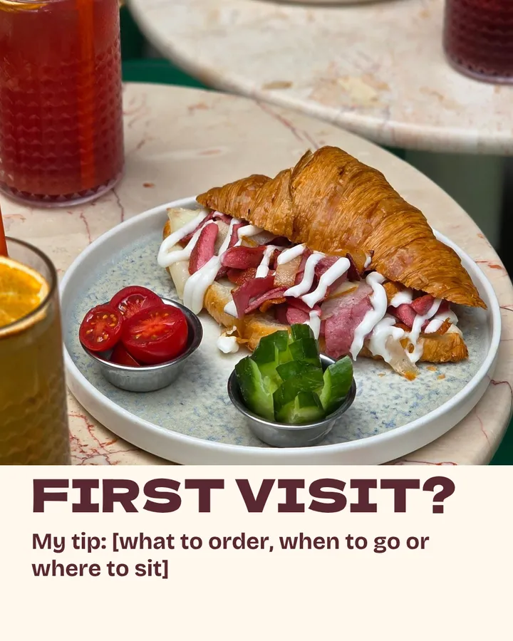 First visit? A croissant sandwich with cured meat and a drizzle of sauce on a café table, with tomatoes, cucumbers and iced tea beside it. My tip: what to order, when to go or where to sit.